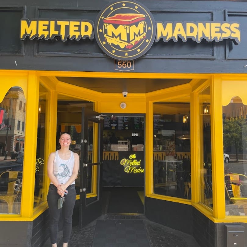 A woman stands smiling in front of The Melted Madness restaurant with a bright yellow facade.