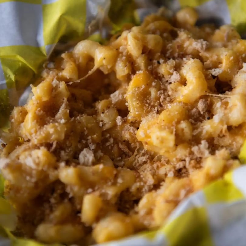 Close-up of creamy mac and cheese with breadcrumbs in a yellow and white checkered paper container.