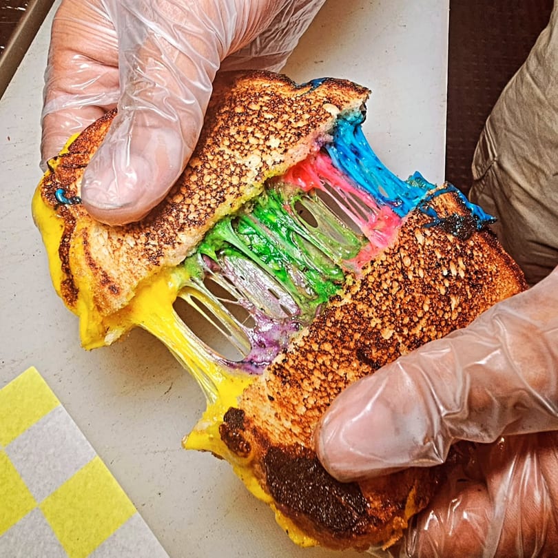 Gloved hands pull apart a grilled cheese sandwich, revealing vibrant rainbow-colored melted cheese inside.