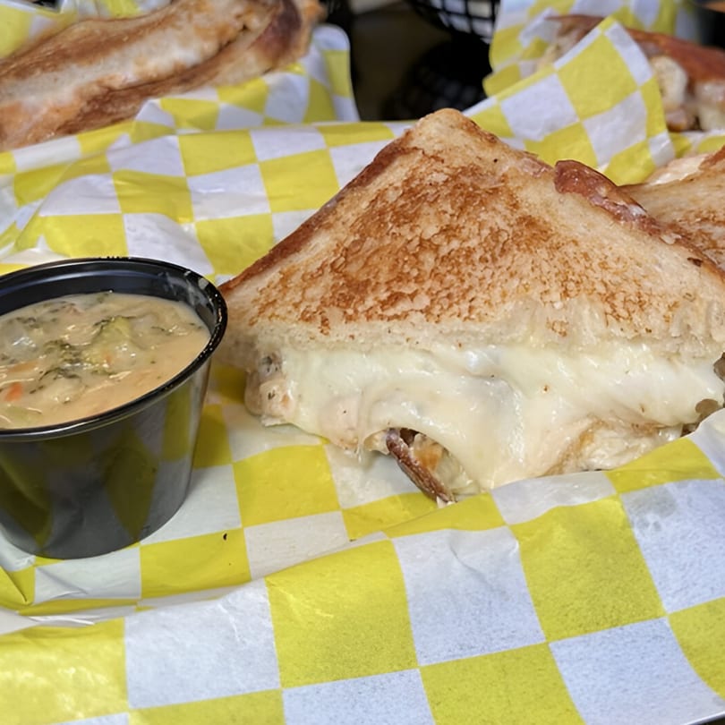 A grilled cheese sandwich with melted cheese and a side of vegetable soup in a checkered basket.