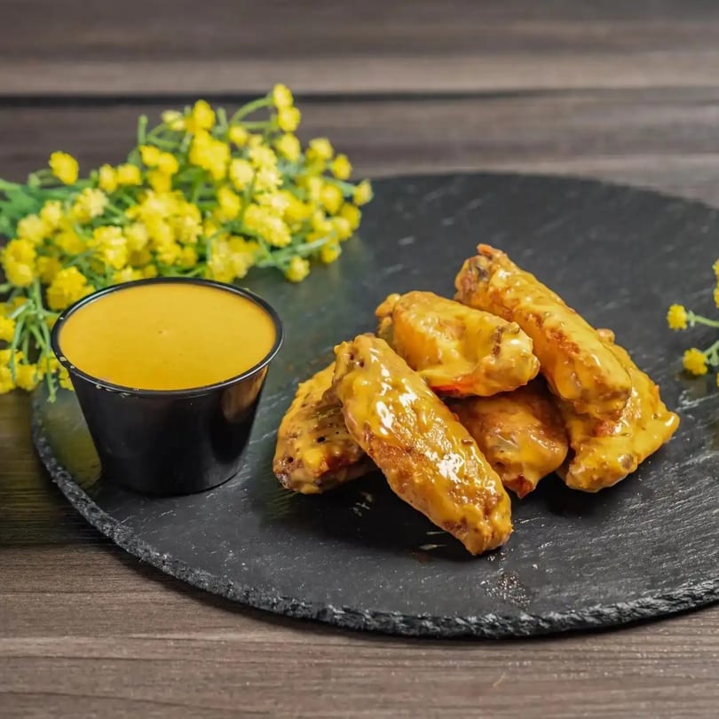 Chicken wings coated in a golden sauce, served with a side of dipping sauce and yellow flowers.