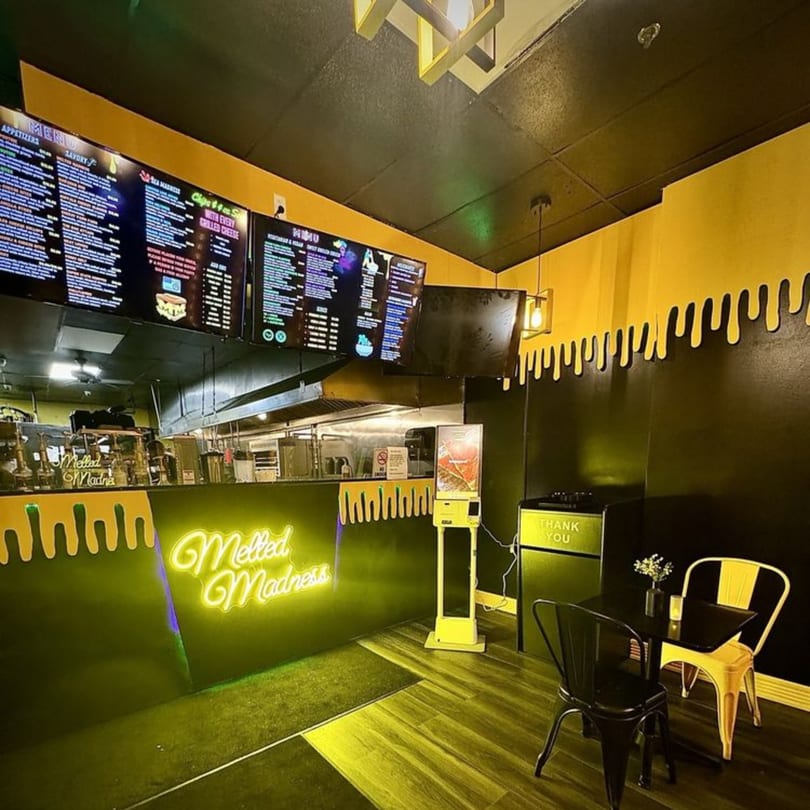 The Melted Madness restaurant interior with black and yellow walls, a neon sign, and menu screens.