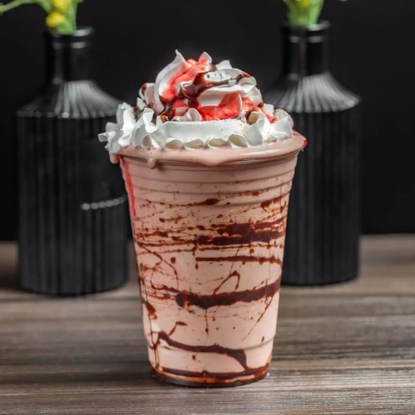 A strawberry milkshake with whipped cream, chocolate, and strawberry syrup in a clear cup.